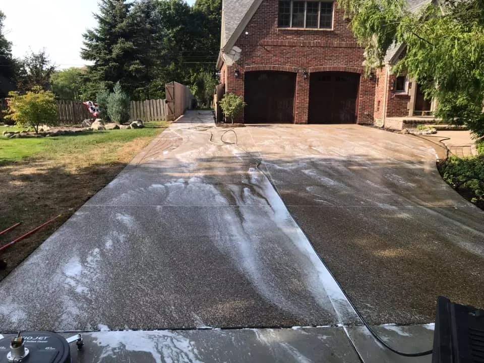 A G&L surface cleaner pass cutting a clean stripe through a dirty concrete driveway in front of a brick garage