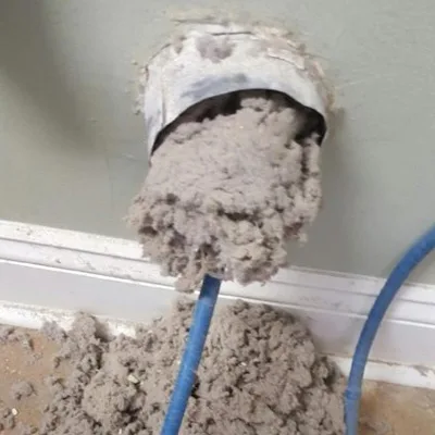 A dryer vent opening packed with gray lint, with more lint pulled out onto the floor below